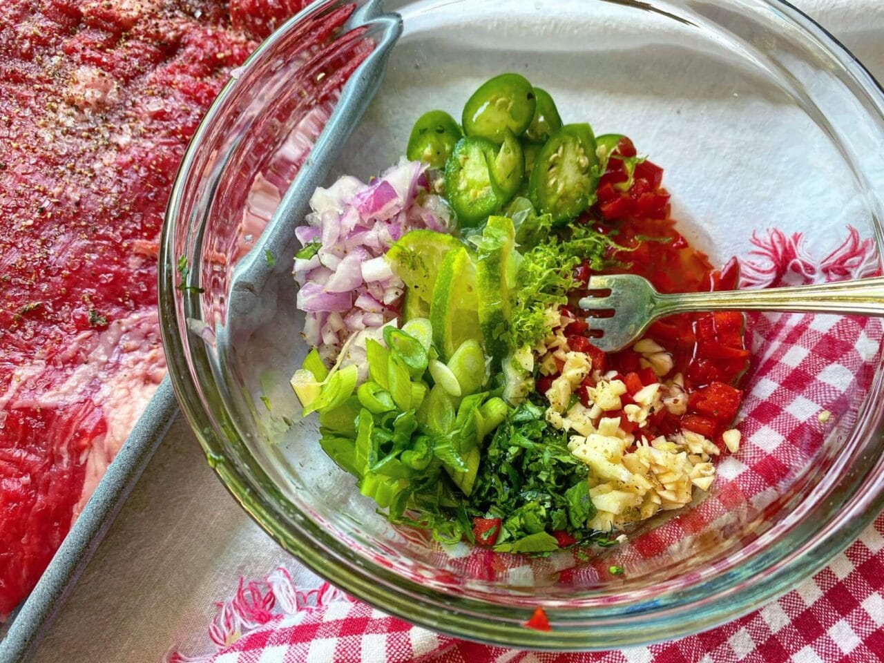 A glass bowl with chopped onions, jalapeños, lime wedges, green onions, cilantro, red peppers, and garlic next to a marinated piece of raw meat.