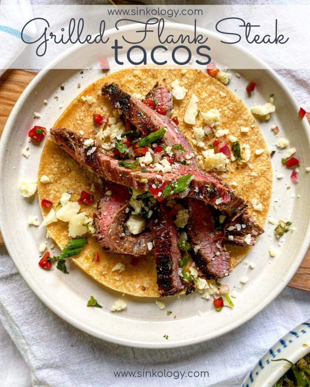 A taco with grilled flank steak, cilantro, red peppers, and crumbled cheese on a corn tortilla, with "Grilled Flank Steak Tacos" and "www.sinkology.com" text above and below the taco.