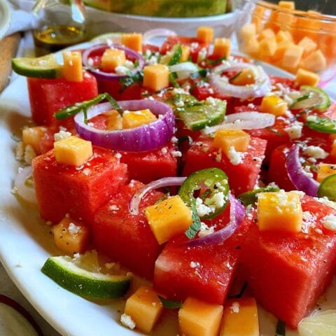 Plate of watermelon cubes topped with cheese, red onion, and jalapeno slices, with additional fruit and a small bottle of dressing in the background.