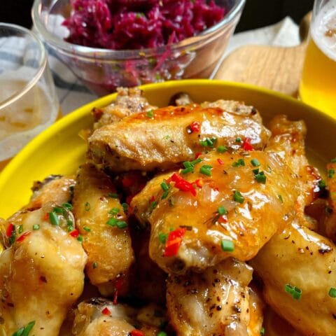 A plate of glazed chicken wings garnished with chopped herbs and red chilies, accompanied by glasses of beer and a bowl of pickled red cabbage.