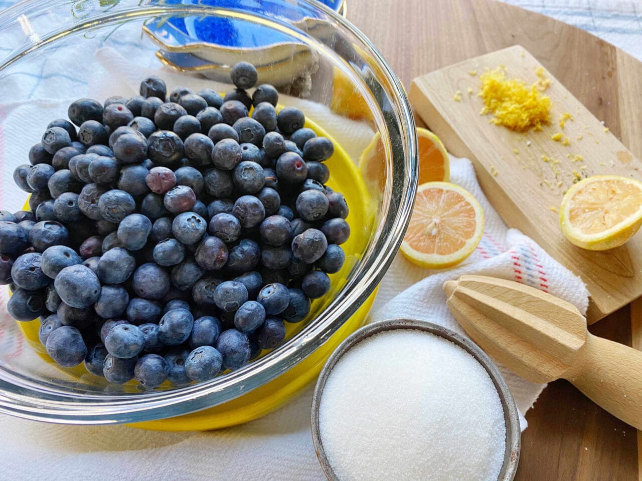 A bowl of fresh blueberries next to a bowl of sugar, halved lemons, lemon zest on a cutting board, and a wooden lemon reamer on a wooden surface.