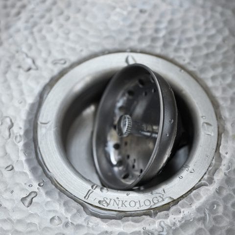 Hammered stainless steel sink bottom with a stainless steel basket strainer drain featured with water droplets.