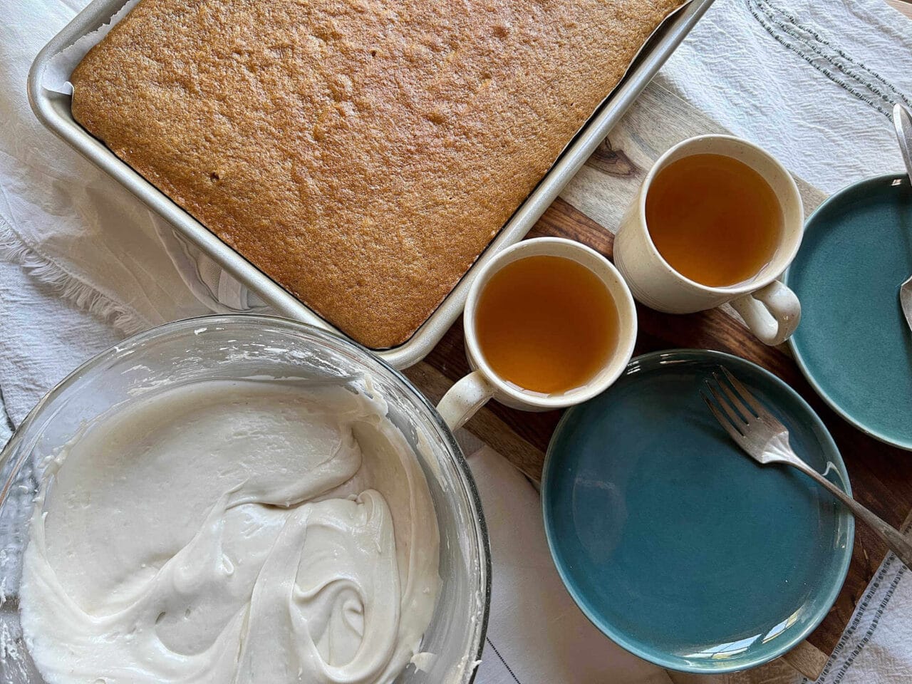 A baked cake in a pan, a bowl of frosting, two cups of tea, and blue plates with forks on a table.