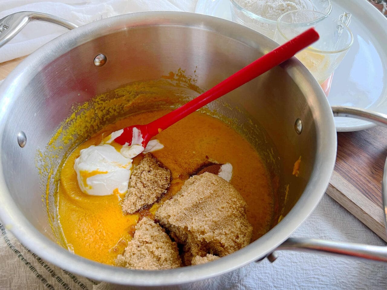 A pot containing pumpkin puree, brown sugar, and sour cream being mixed with a red spatula.