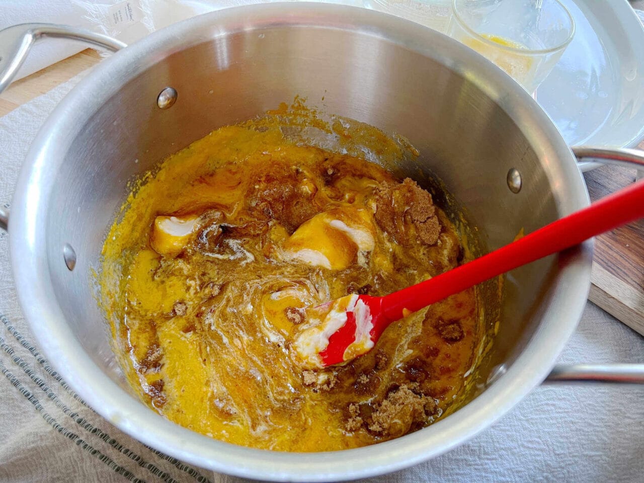 Ingredients, including pumpkin puree, brown sugar, and sour cream, being mixed in a pot with a red spatula.