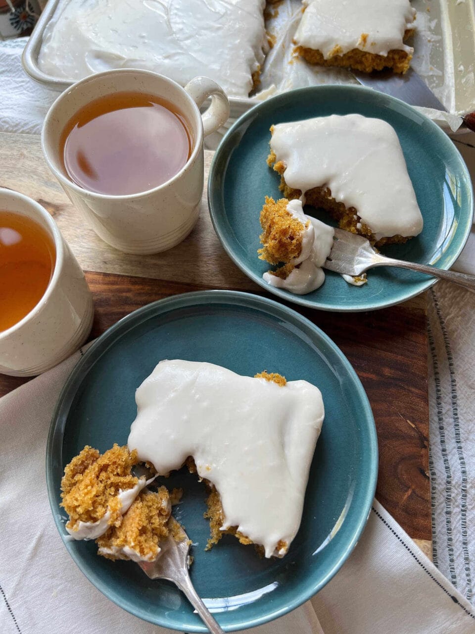 Two plates with slices of frosted cake and forks, next to cups of tea, with the remaining cake in the background.