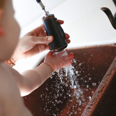 A double bowl copper kitchen sink with an adult hand spraying water from a bronze pull down faucet onto the hand of a baby's outstretched arm.