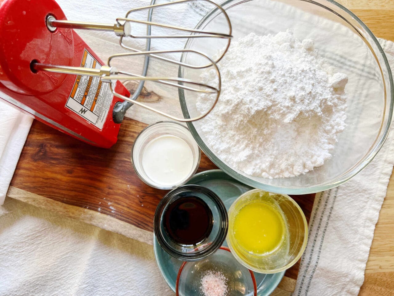 Ingredients for maple buttercream frosting, including powdered sugar, cream, vanilla extract, melted butter, and salt, arranged next to a red hand mixer.