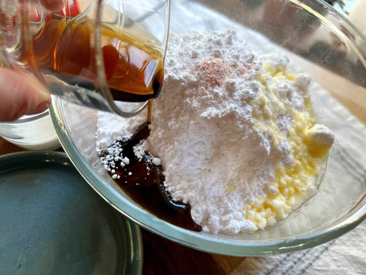 Maple syrup being poured into a bowl containing powdered sugar, melted butter, and salt for making maple buttercream frosting.