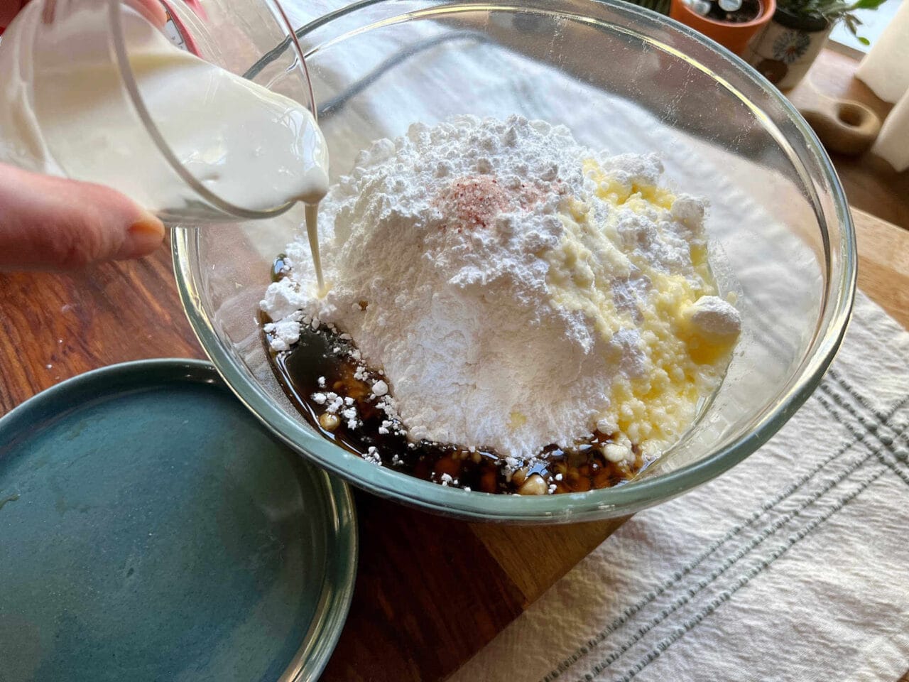 Milk being poured into a bowl containing powdered sugar, maple syrup, melted butter, and salt for making maple buttercream frosting.
