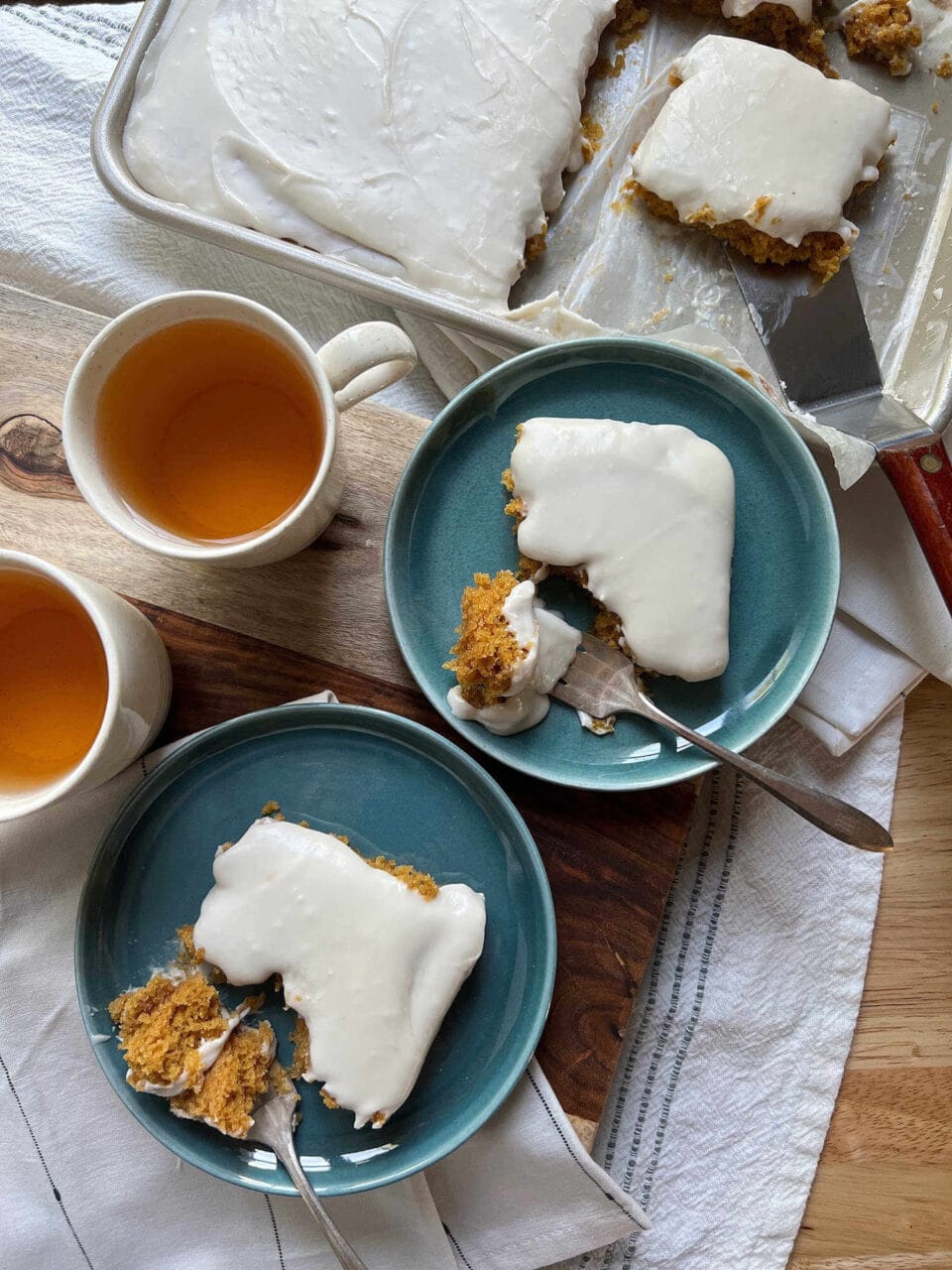 Two plates with frosted cake slices and forks next to cups of tea, with the remaining frosted cake in the background.