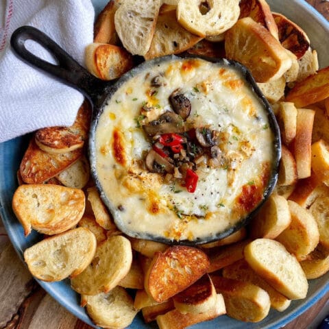 A cast iron skillet filled with cheesy dip topped with mushrooms and chopped red peppers, surrounded by toasted bread slices on a blue plate.