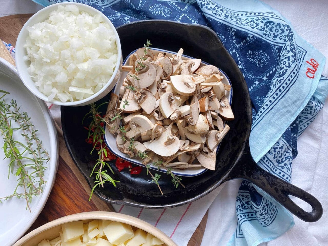 A cast iron skillet containing a bowl of sliced mushrooms, fresh herbs, and red chili slices, with a separate bowl of chopped onions beside it.