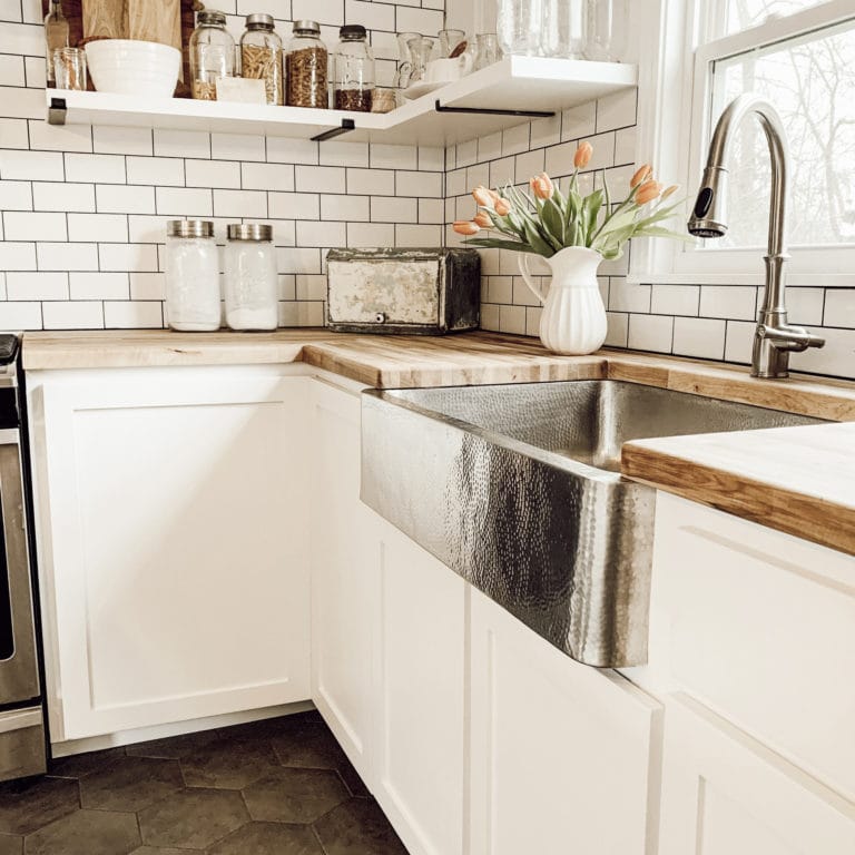 A hand hammered stainless steel undermount farmhouse kitchen sink with butcher block countertops, white cabinets and subway tile backsplash.