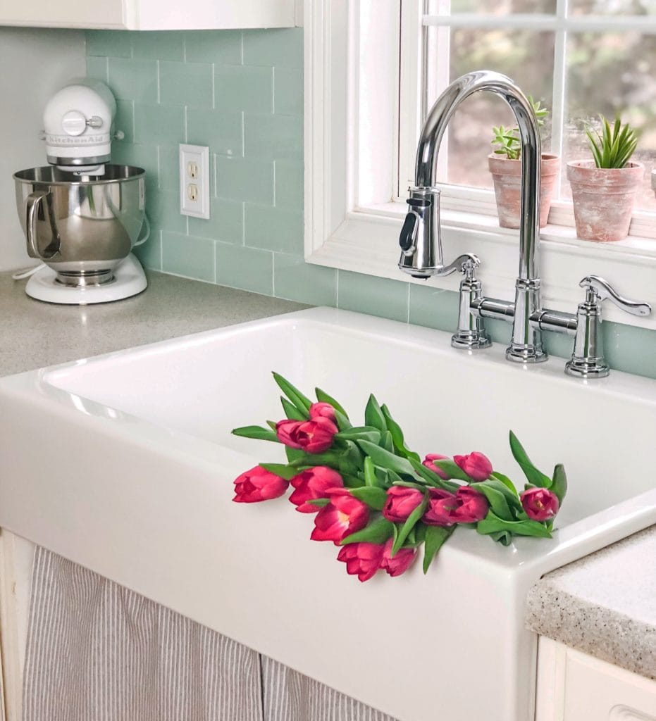 A glossy white rectangular Quick-Fit® drop-in farmhouse sink with teal tile backsplash and pink tulip flowers in the single basin.