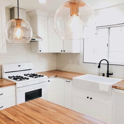 A glossy white rectangular Quick-Fit® drop-in farmhouse sink, dark metal faucet set in butcher block countertops in a bright white kitchen with a large window and island.