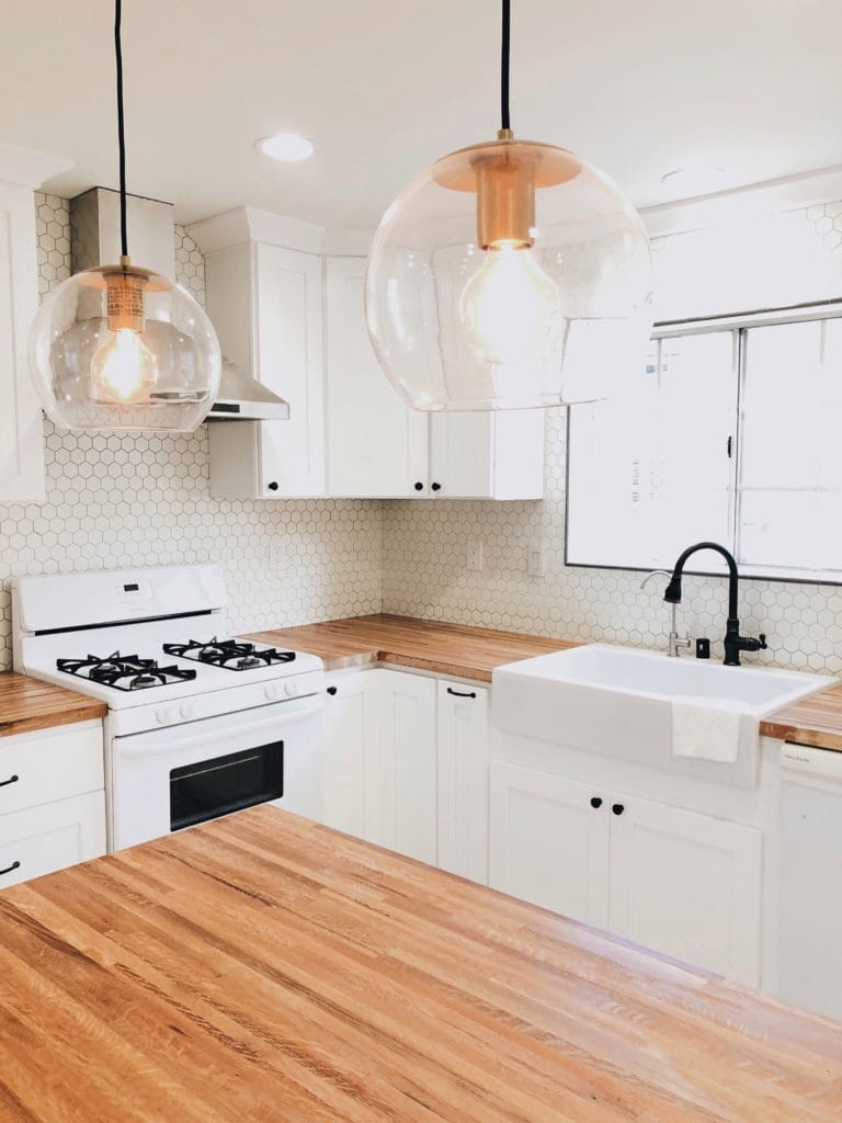 A glossy white rectangular Quick-Fit® drop-in farmhouse sink, dark metal faucet set in butcher block countertops in a bright white kitchen with a large window and island.