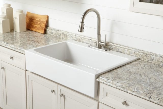 A glossy white rectangular Quick-Fit® drop-in farmhouse sink made in fireclay with a brushed nickel faucet, marble countertops and tan cabinets.