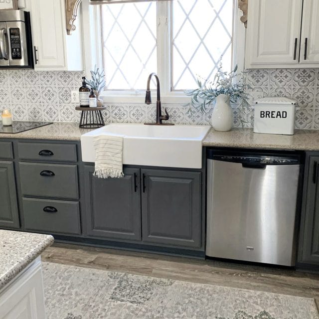 A glossy white rectangular Quick-Fit® drop-in farmhouse sink made in fireclay, a bronze faucet and drain set in gray coutertops and cabinets with a window above the sink.
