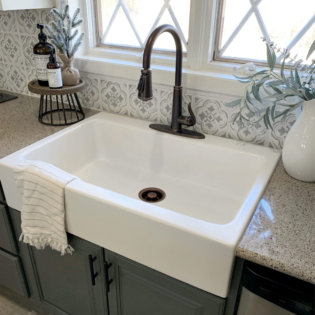 A glossy white rectangular Quick-Fit® drop-in farmhouse sink made in fireclay, a bronze faucet and drain set in gray coutertops and cabinets with a window above the sink.