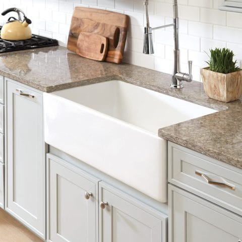 A kitchen with a white farmhouse sink, granite countertops, and light gray cabinets. A cutting board and a potted plant sit next to a stainless steel faucet.