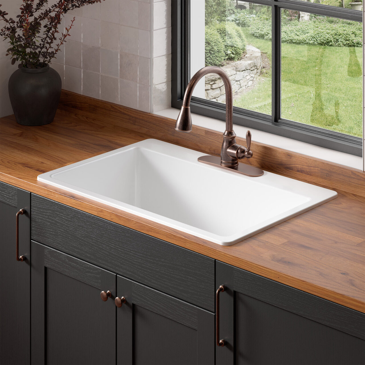A white fireclay drop-in sink with bronze faucet in butcher block countertops with dark gray cabinets in front of a window