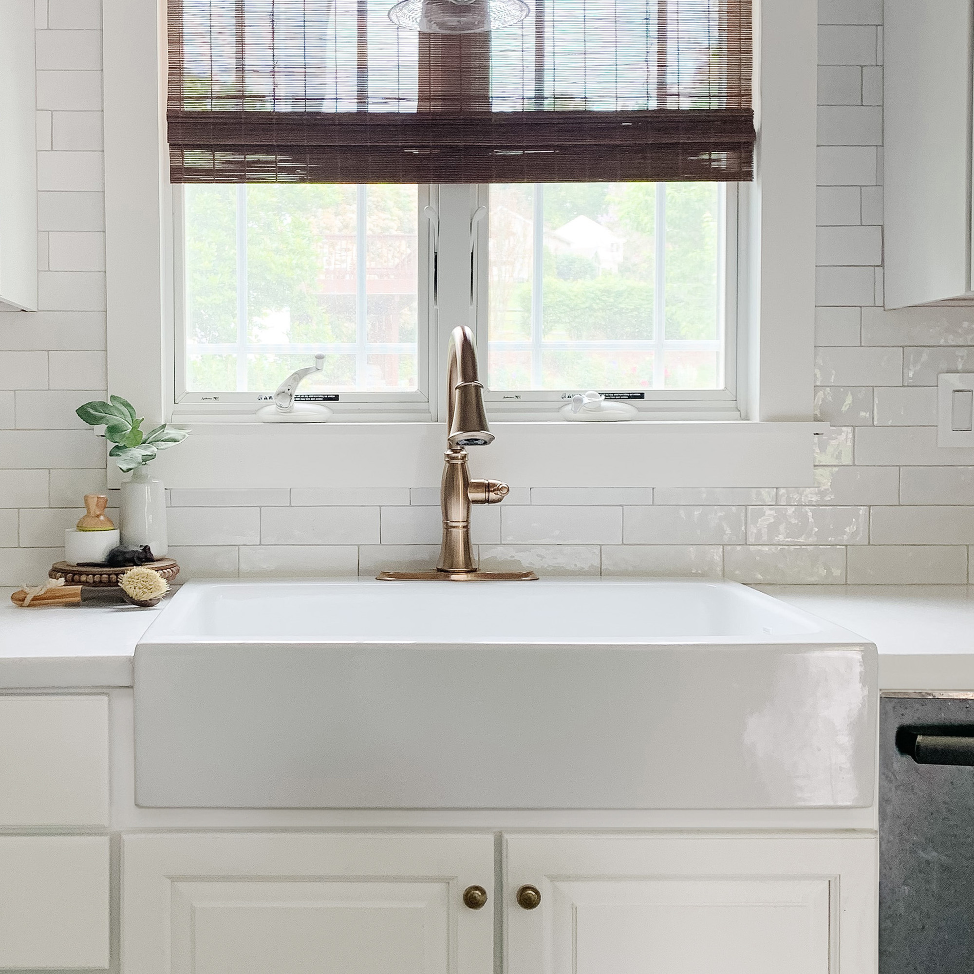 A glossy white rectangular Quick-Fit® drop-in farmhouse sink made in fireclay with a traditional faucet, set in white countertop with white cabinets in front of large window with wooden shades.