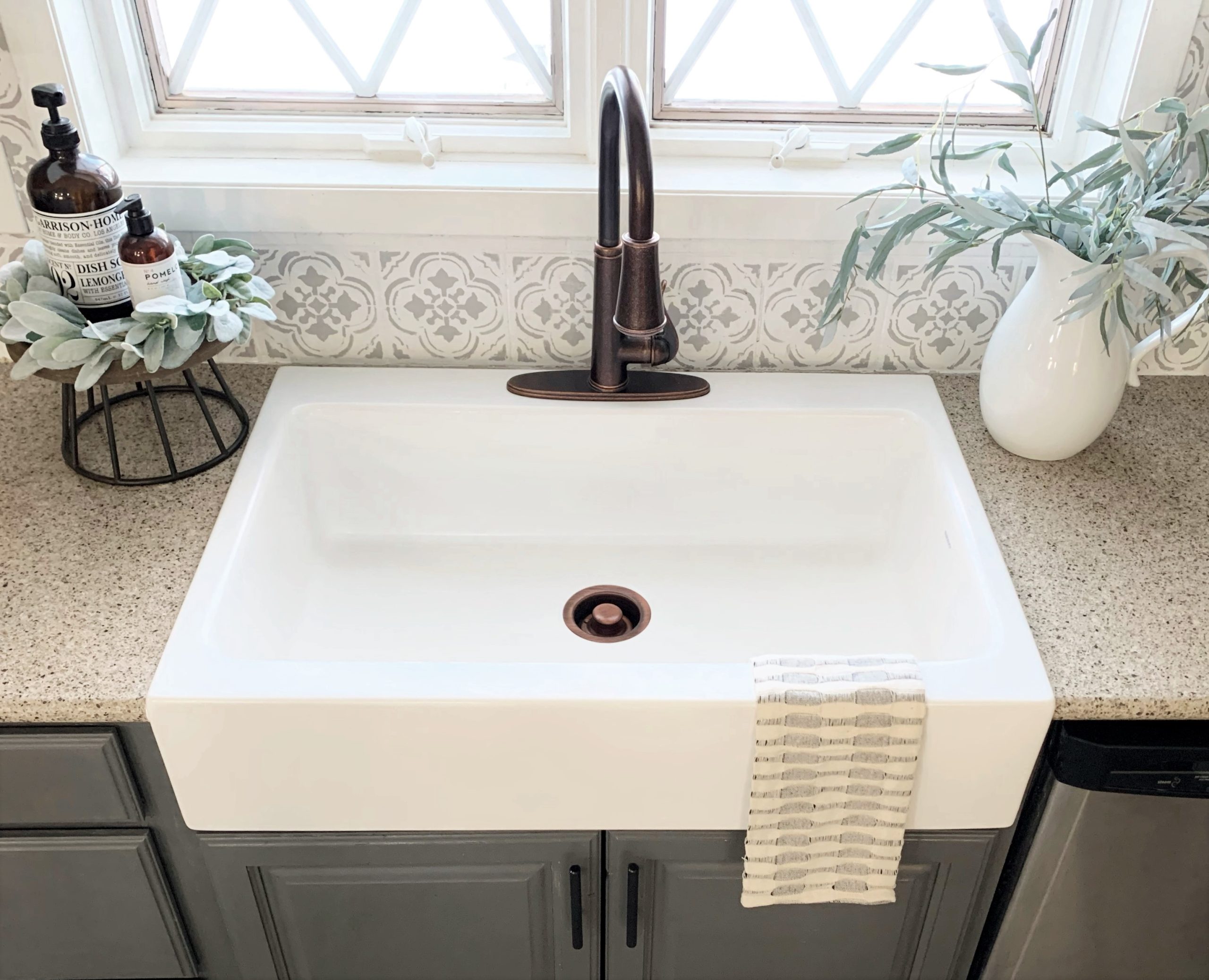 A matte white rectangular Quick-Fit® drop-in farmhouse sink made in fireclay with a bronze faucet, gray cabinets and tan countertop with large window.