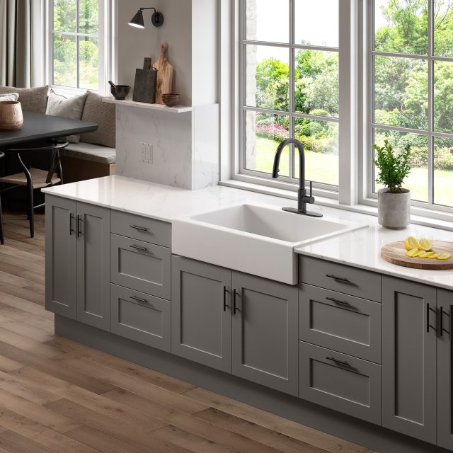 A glossy white rectangular Quick-Fit® drop-in farmhouse sink made in fireclay with a modern matte black faucet, white countertop and gray cabinets with a large window overlooking a lawn.