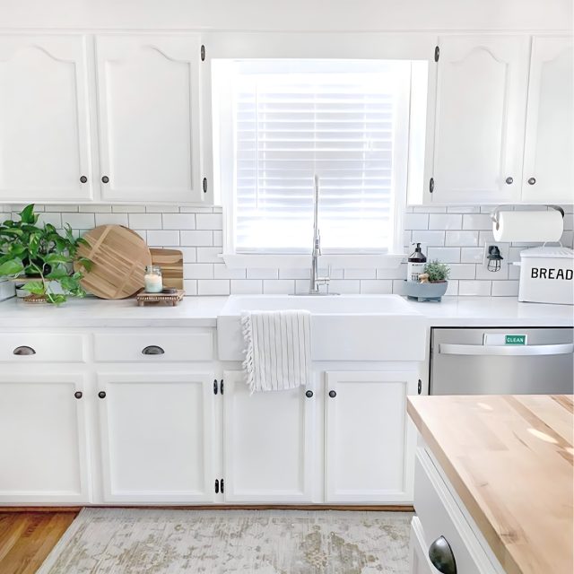 A glossy white rectangular Quick-Fit® drop-in farmhouse sink made in fireclay with a chrome faucet set in a white countertop and cabinets under a window.