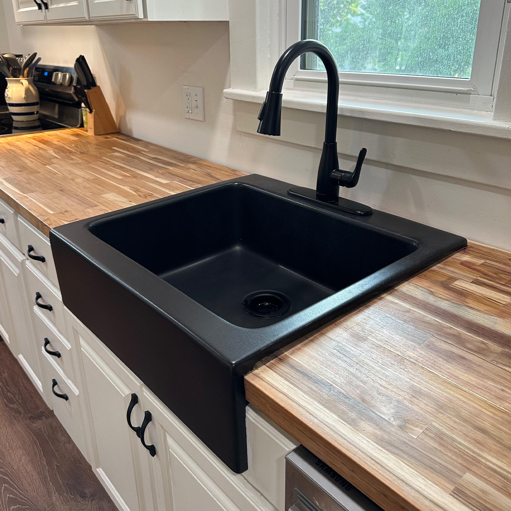 A kitchen with a matte black rectangular Quick-Fit® drop-in farmhouse sink made in fireclay with a black faucet, butcher block countertops and white cabinets.