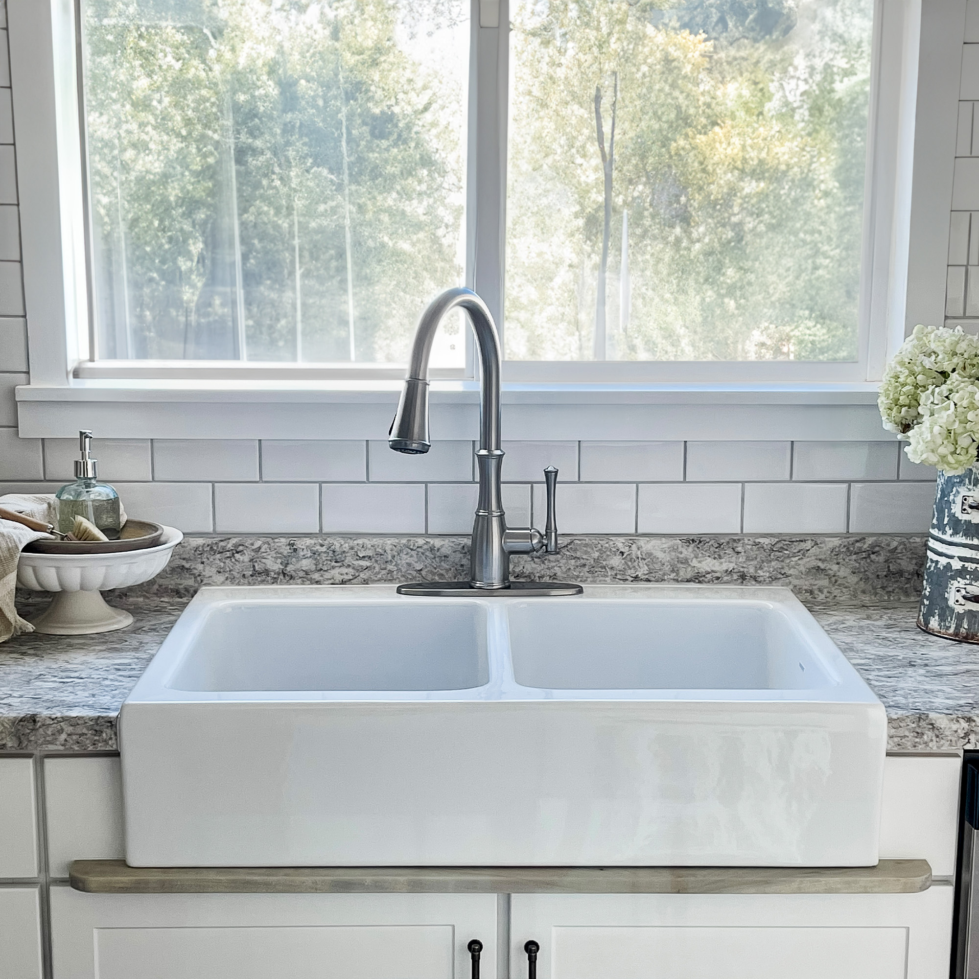 A glossy white rectangular Quick-Fit® double bowl drop-in farmhouse sink made in fireclay with a brushed stainless faucet in front of a large window.