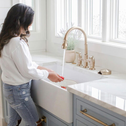 A woman washes vegetables at a white farmhouse sink with a brass faucet. The kitchen features a light-colored marble countertop and blue cabinetry, with a potted plant on the windowsill above the sink.