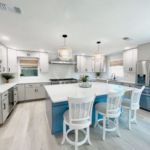 A modern kitchen with a blue island, white cabinets, and stainless steel appliances. White barstools and pendant lights complete the space.