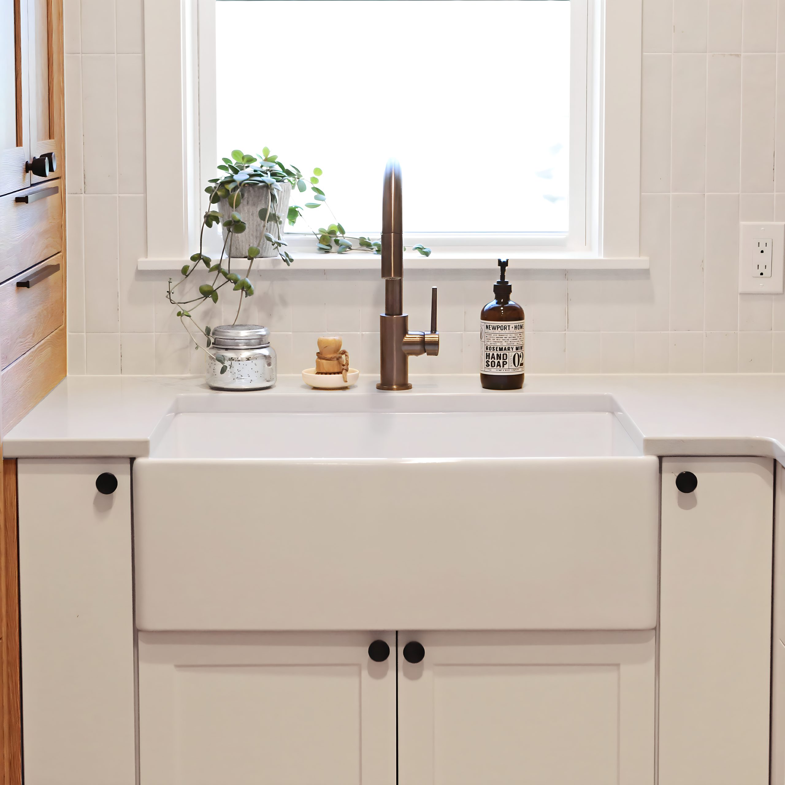A kitchen with a matte white rectangular undermount farmhouse sink made in fireclay, in front of a window, set in a solid surface white countertop with a modern pull-down faucet and white cabinets.