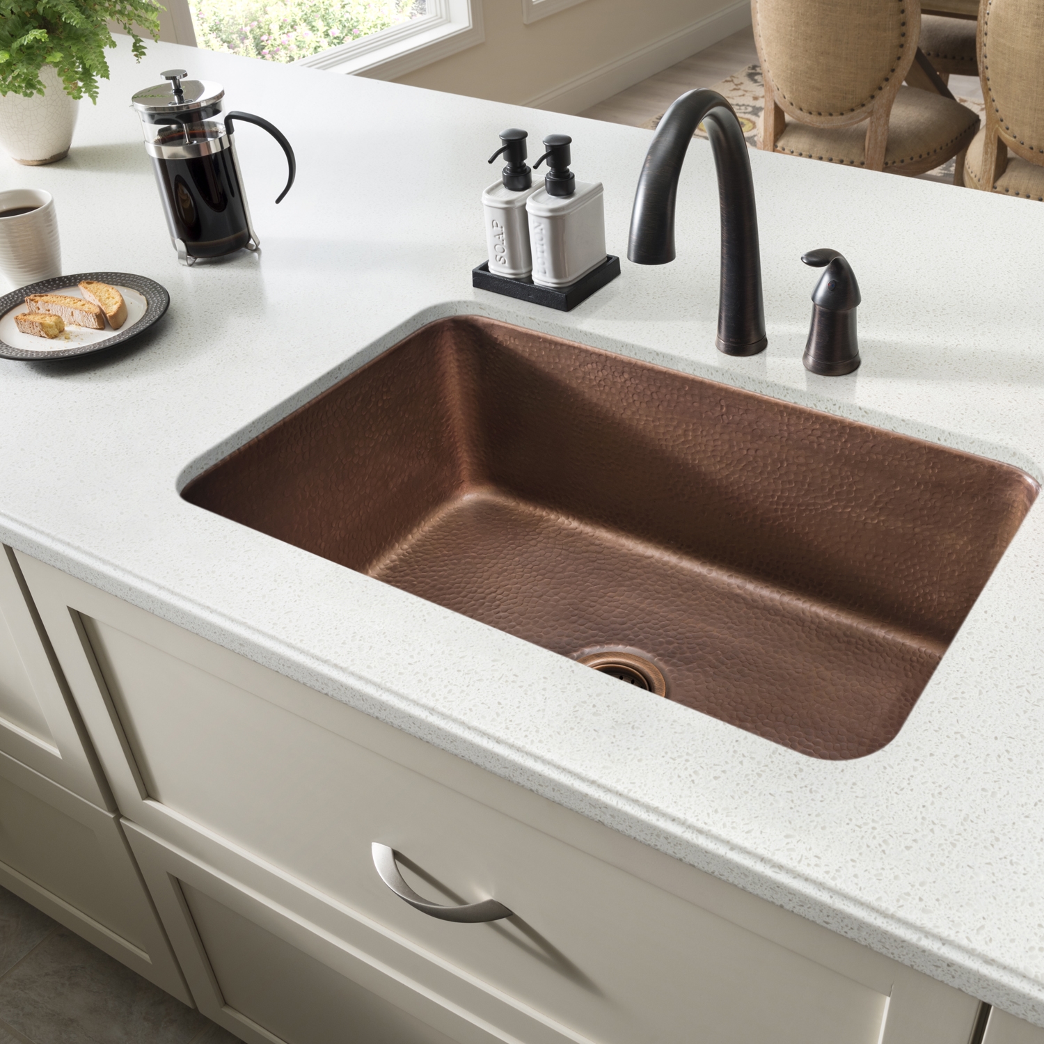 A kitchen with a copper undermount sink, bronze faucet, white countertop and cabinets.