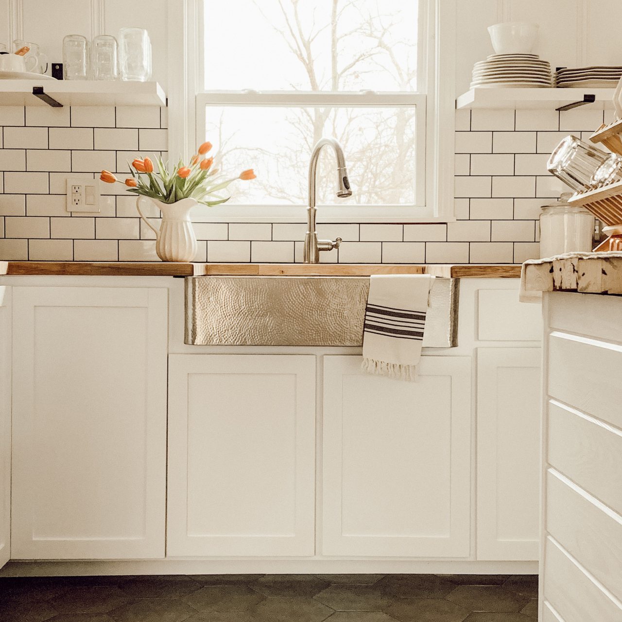 Kitchen with crafted stainless steel apron front farmhouse sink set in white cabinets with butcherblock countertop under large window.