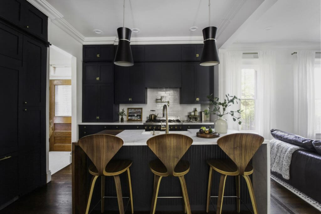 A modern kitchen with dark cabinets, a white marble island with a sink, three wooden barstools, and pendant lights overhead, opening into a bright living area.