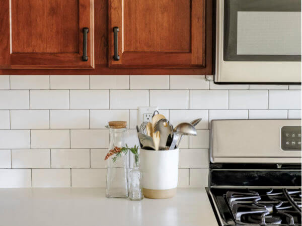 White kitchen counter with cooking utensils in a white holder and glass bottles beside a stainless steel stove.