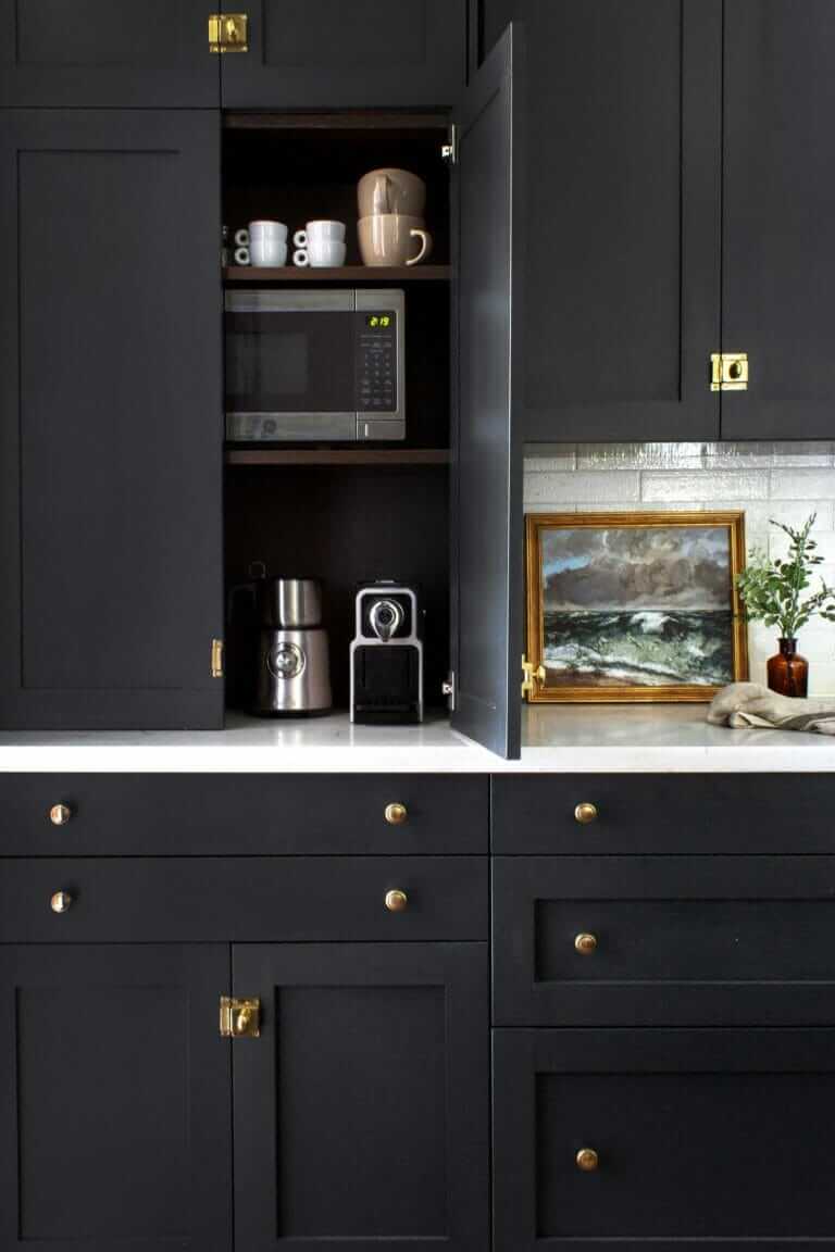 A kitchen with dark cabinets, one of which is open to reveal shelves with a microwave, coffee maker, and mugs; a white countertop features a small framed painting and a potted plant.