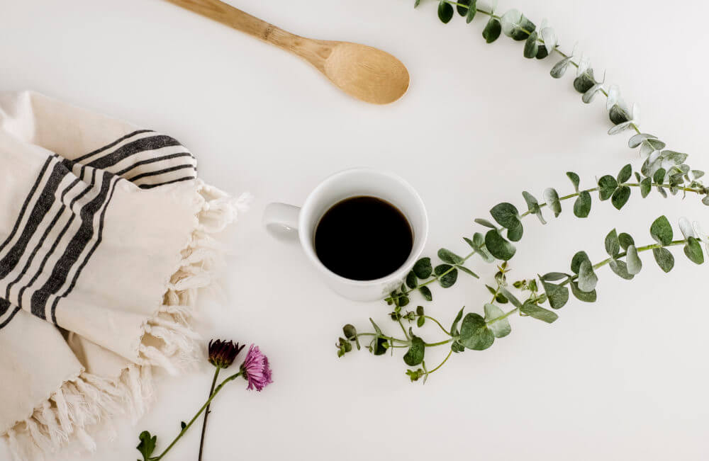 A flat lay of a cozy setup featuring a wooden spoon, a white coffee cup filled with black coffee, a striped fringed cloth, a purple flower, and green leaves arranged on a white surface.