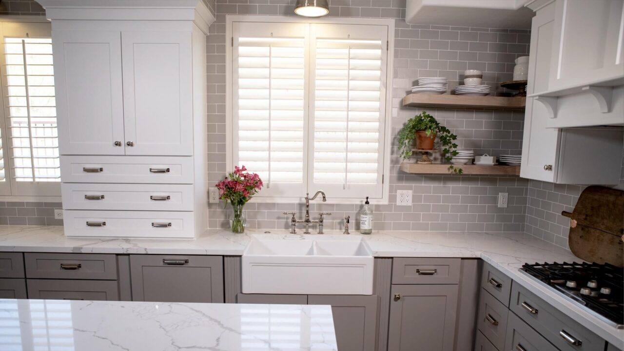 Traditional kitchen with white fireclay farmhouse sink, gray cabinets and light decor on open shelves.