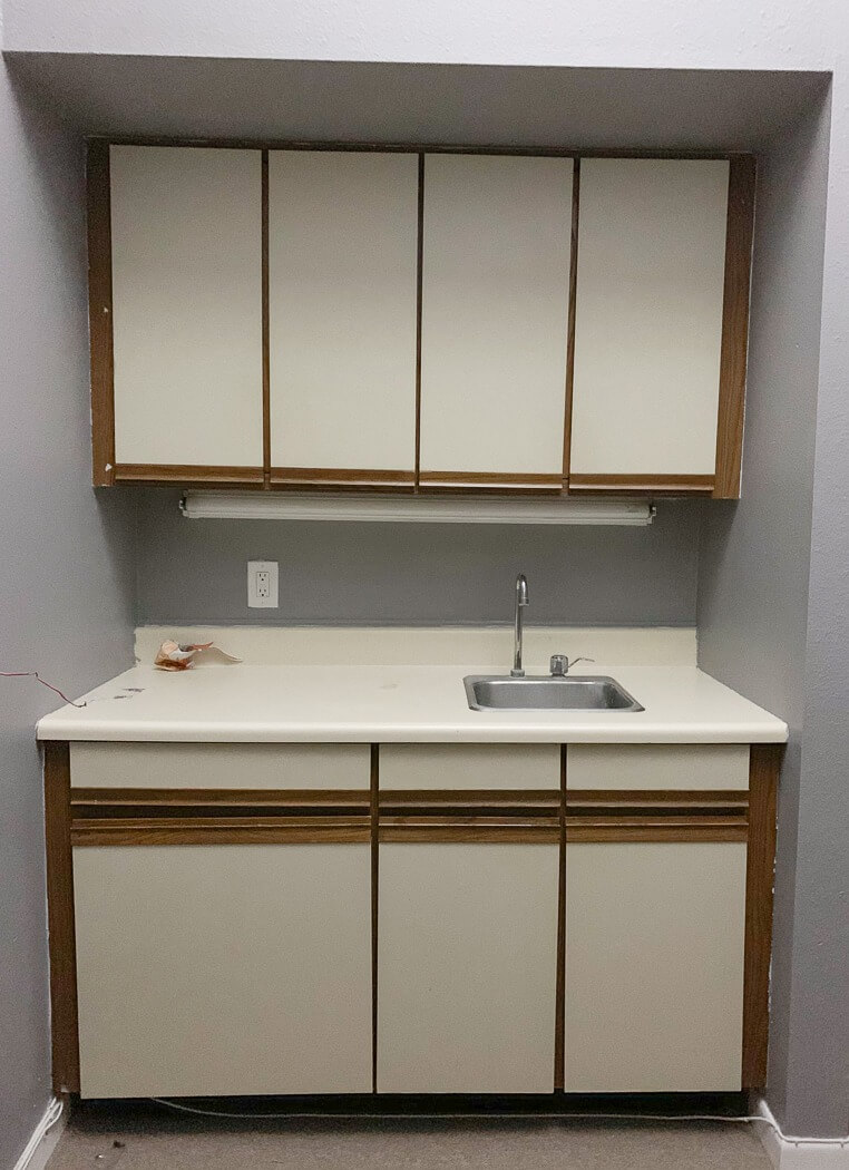 Simple kitchen setup with beige cabinets, a small sink, and a plain countertop against a gray wall.