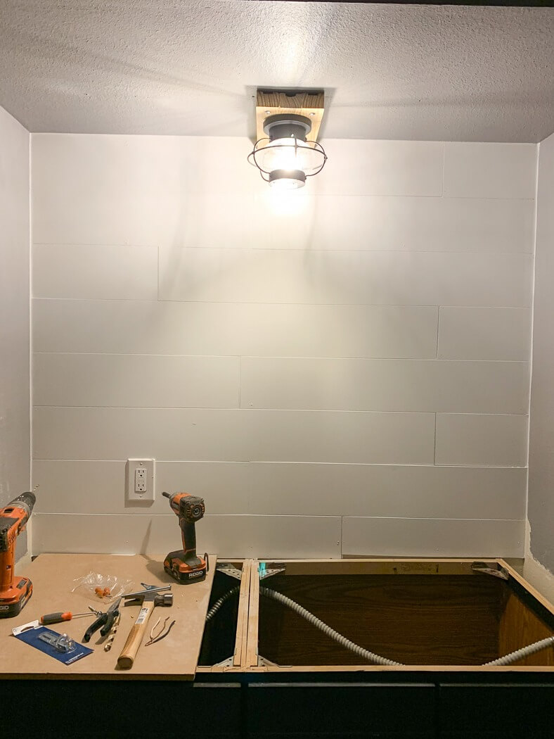 Work area illuminated by a new light fixture, with tools and drills on a partially constructed countertop against a shiplap wall.