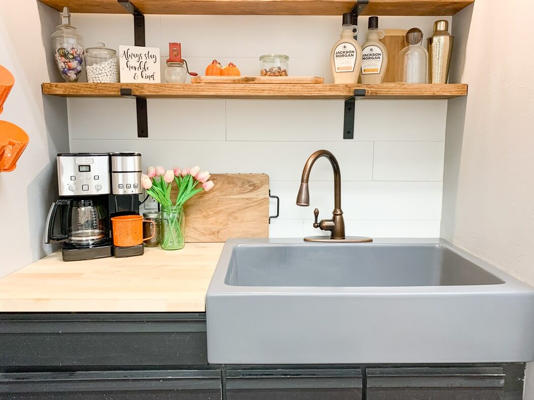 Cozy kitchen nook with a gray sink, bronze faucet, coffee maker, wooden shelves holding decor and bottles, and a vase of pink tulips on the counter.