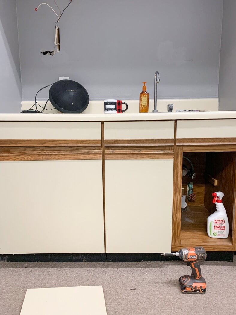 Partially disassembled kitchen cabinet with tools and cleaning supplies, wires exposed above the countertop, and a drill on the floor.