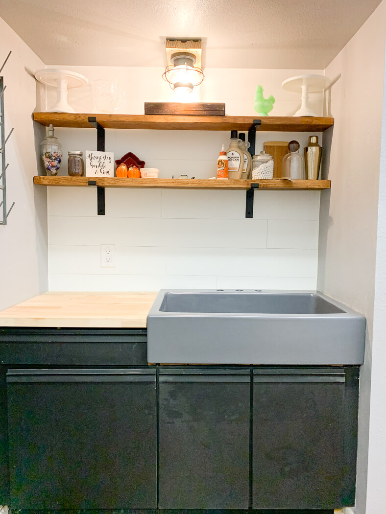 Small kitchen area with dark cabinets, gray sink, wooden shelves displaying decor items, and a light fixture overhead.