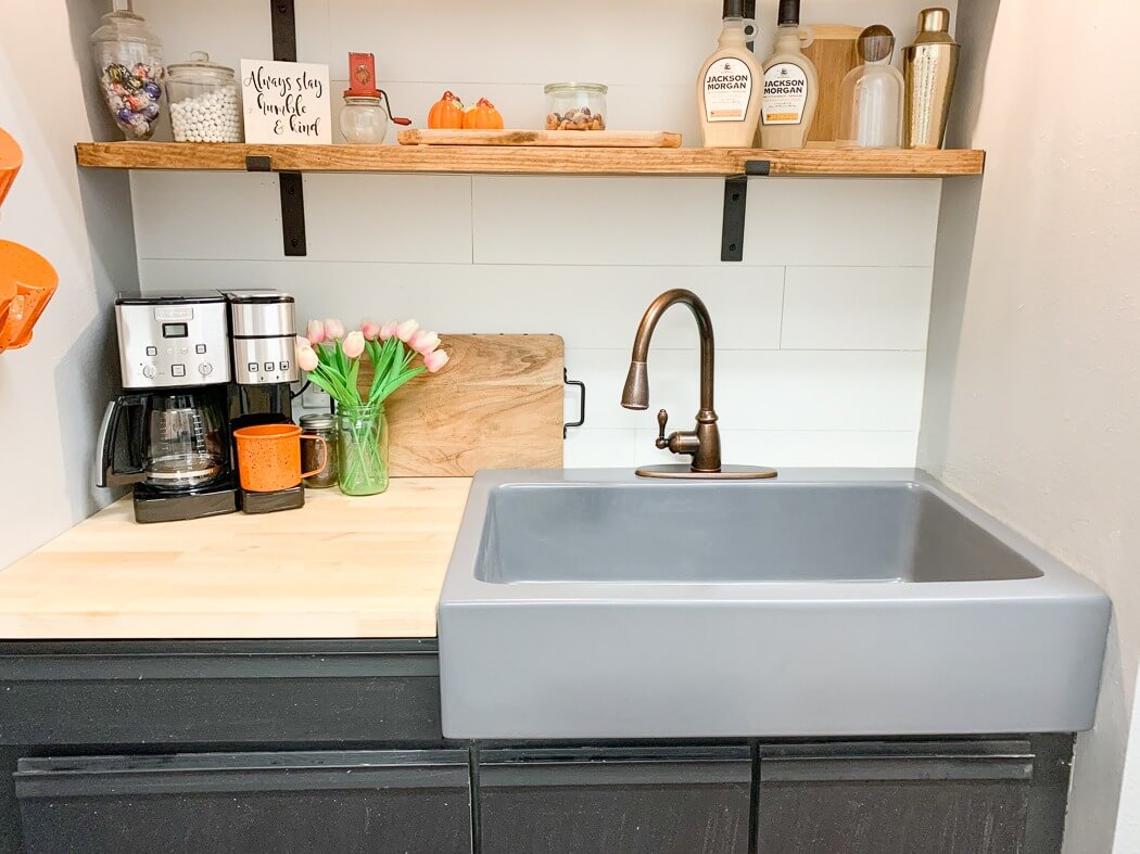 Cozy kitchen corner featuring a gray sink with a bronze faucet, a coffee maker with an orange mug, a vase of pink tulips, and wooden shelves displaying decor items above.