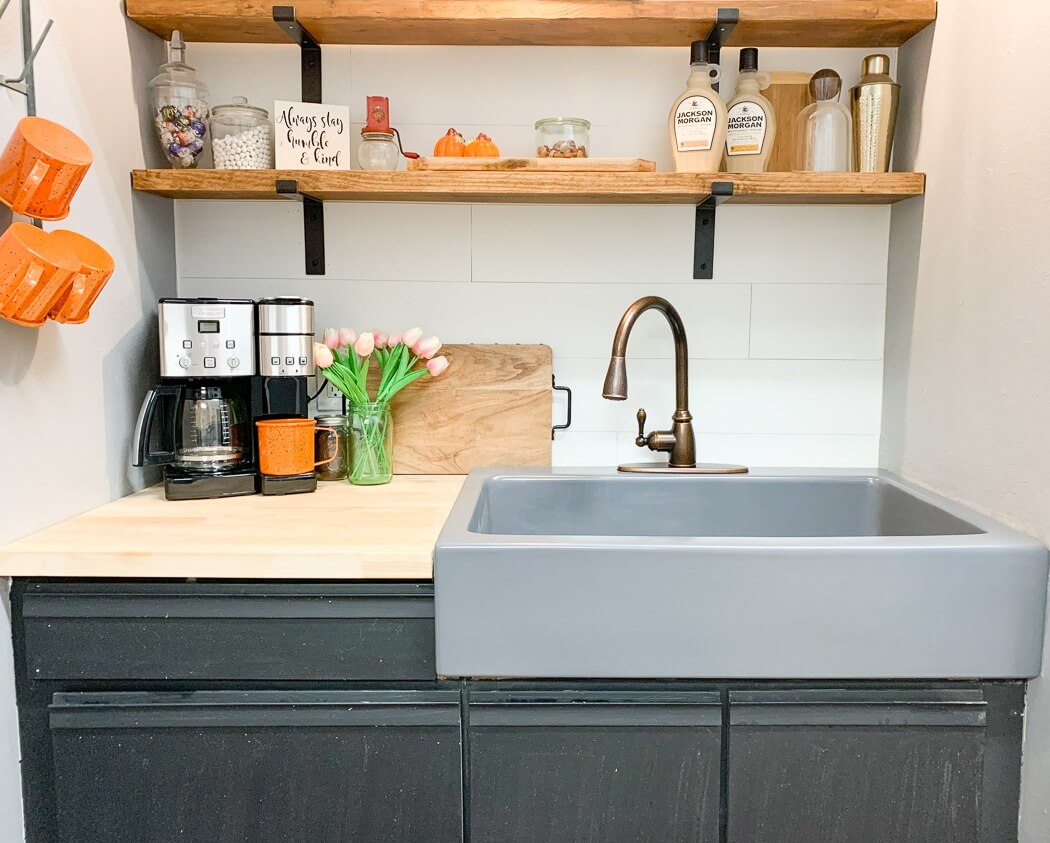 Stylish kitchen setup with a gray sink, bronze faucet, coffee maker, orange mugs, and wooden shelves holding decor and bottles, along with a vase of pink tulips on the counter.