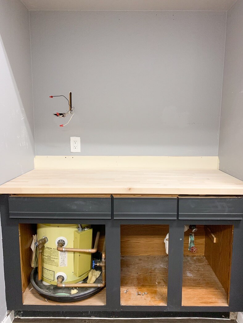 Kitchen cabinet under construction with exposed pipes, a small water heater, and bare wooden countertop, wires hanging from the wall.